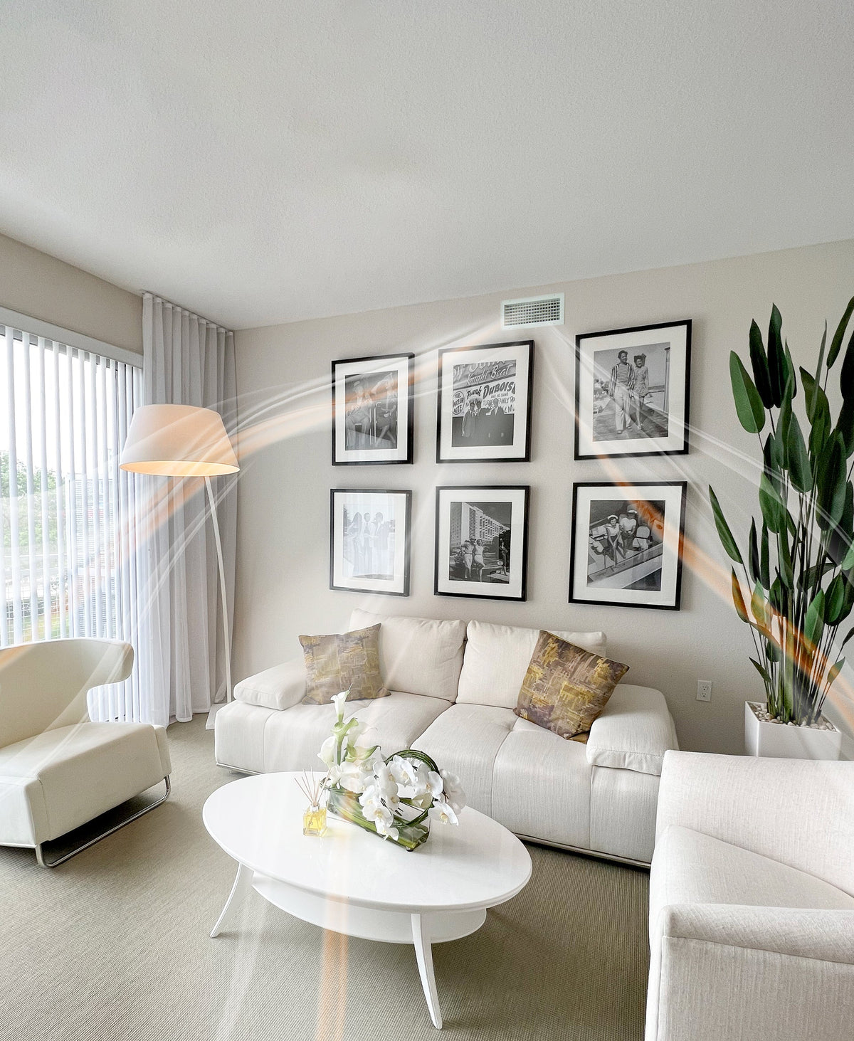 Bright and modern living room with a white sofa set, black-and-white framed artwork, a tall green plant, and a reed diffuser with white flowers on a white coffee table. Soft aroma trails are digitally illustrated in the air.