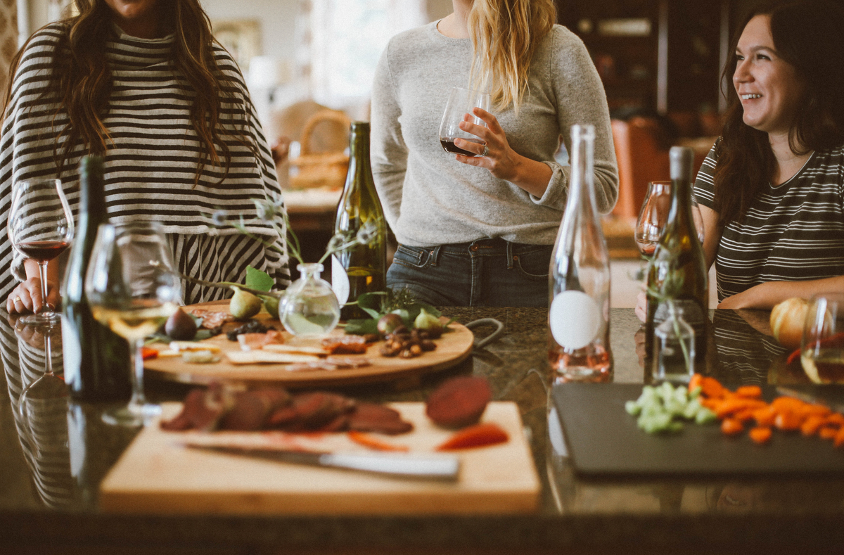 Three women gathered around a kitchen island, enjoying wine and conversation, with charcuterie boards, fresh vegetables, and bottles on the counter creating a warm and inviting atmosphere.