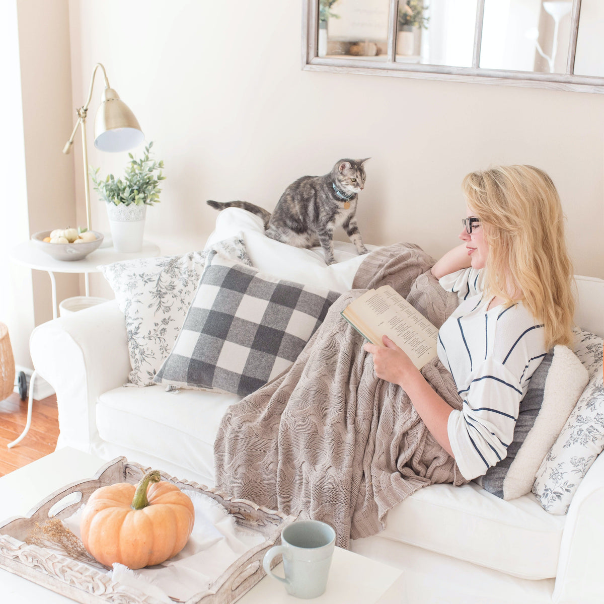 Cozy fall scene with a woman reading on a white sofa, wrapped in a taupe knit blanket, surrounded by soft pillows, with a gray cat nearby and a tray holding a pumpkin and mug on the coffee table.