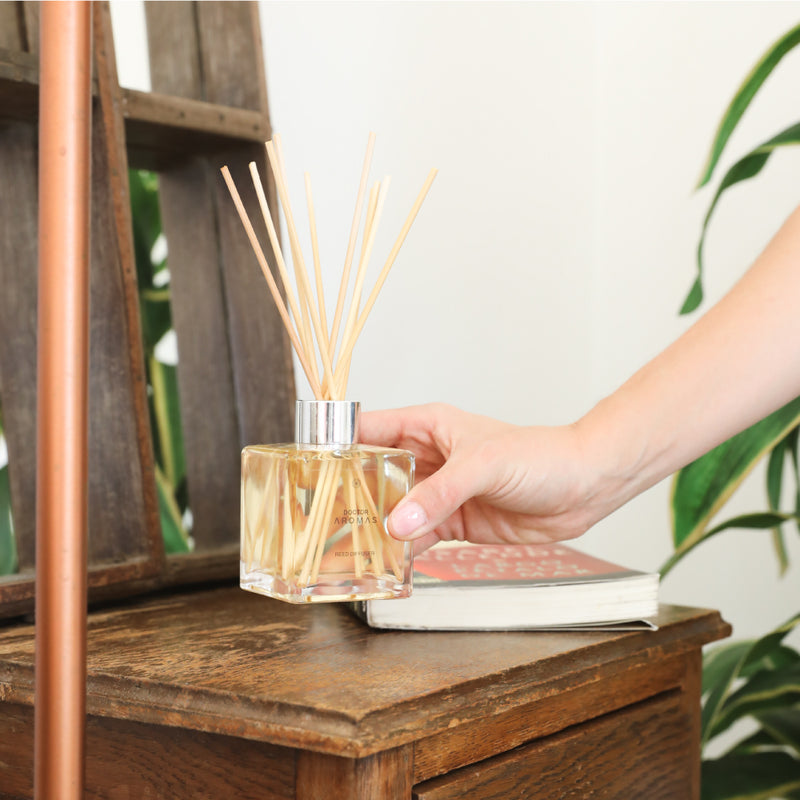 Clear square glass reed diffuser with light-colored oil and natural reeds on rustic wooden surface with hand adjusting reeds. - Oil Reed Diffuser Twin Pack | Holiday - Holiday - Doctor Aromas