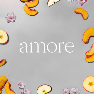 White candle container with "amore" text surrounded by peach slices, apple slices, and pink flower petals on gray background. - Candle | Amore - Amore - Doctor Aromas