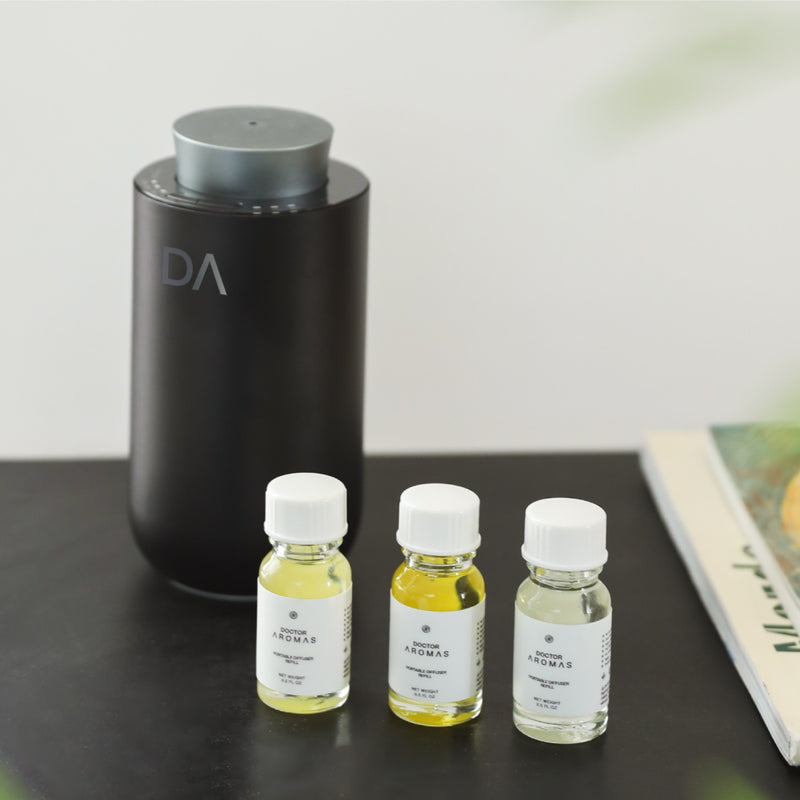 Three small translucent yellow liquid refill bottles with white caps in front of a tall matte black portable diffuser on a. - Portable Diffuser Refills Pack of Three | Signature Collection - Signature Collection - Doctor Aromas