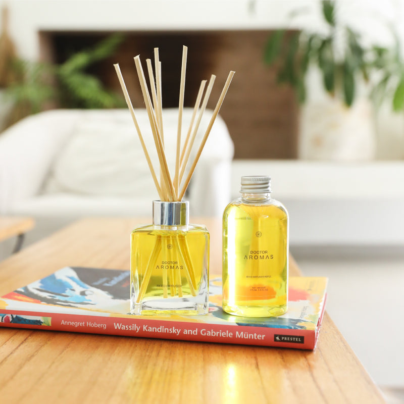 Clear glass reed diffuser with natural wooden sticks and a matching refill bottle on colorful books in a cozy living room. - Oil Reed Diffuser & Refill Bundle | Holiday - Holiday - Doctor Aromas