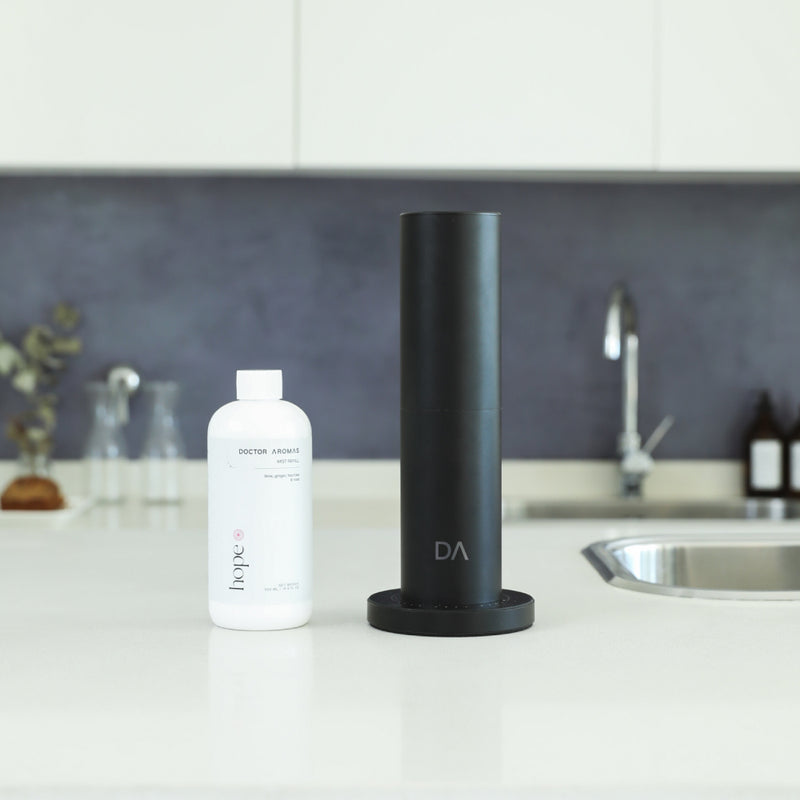 Matte black cylindrical aroma diffuser with a flat round base on white kitchen countertop next to white refill bottle. - Aroma Diffuser 500 | Holiday - Holiday / Black - Doctor Aromas