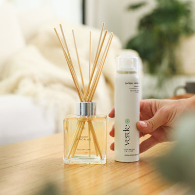 Square glass reed diffuser with natural wooden sticks beside a white spray bottle held by a hand on a wooden table. - Aroma Set | Holiday - Holiday - Doctor Aromas
