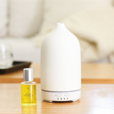 White ceramic ultrasonic diffuser with silver buttons next to small amber glass bottle of yellow diffuser oil on wooden table. - Diffuser Drops | Holiday - Holiday - Doctor Aromas