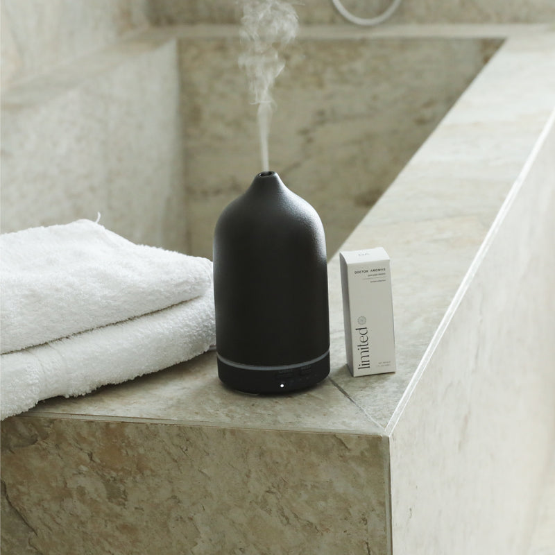 Matte black stone diffuser emitting mist on beige marble ledge next to small white box and folded white towel. - Aromatherapy Stone Diffuser | Blush - Blush / White - Doctor Aromas